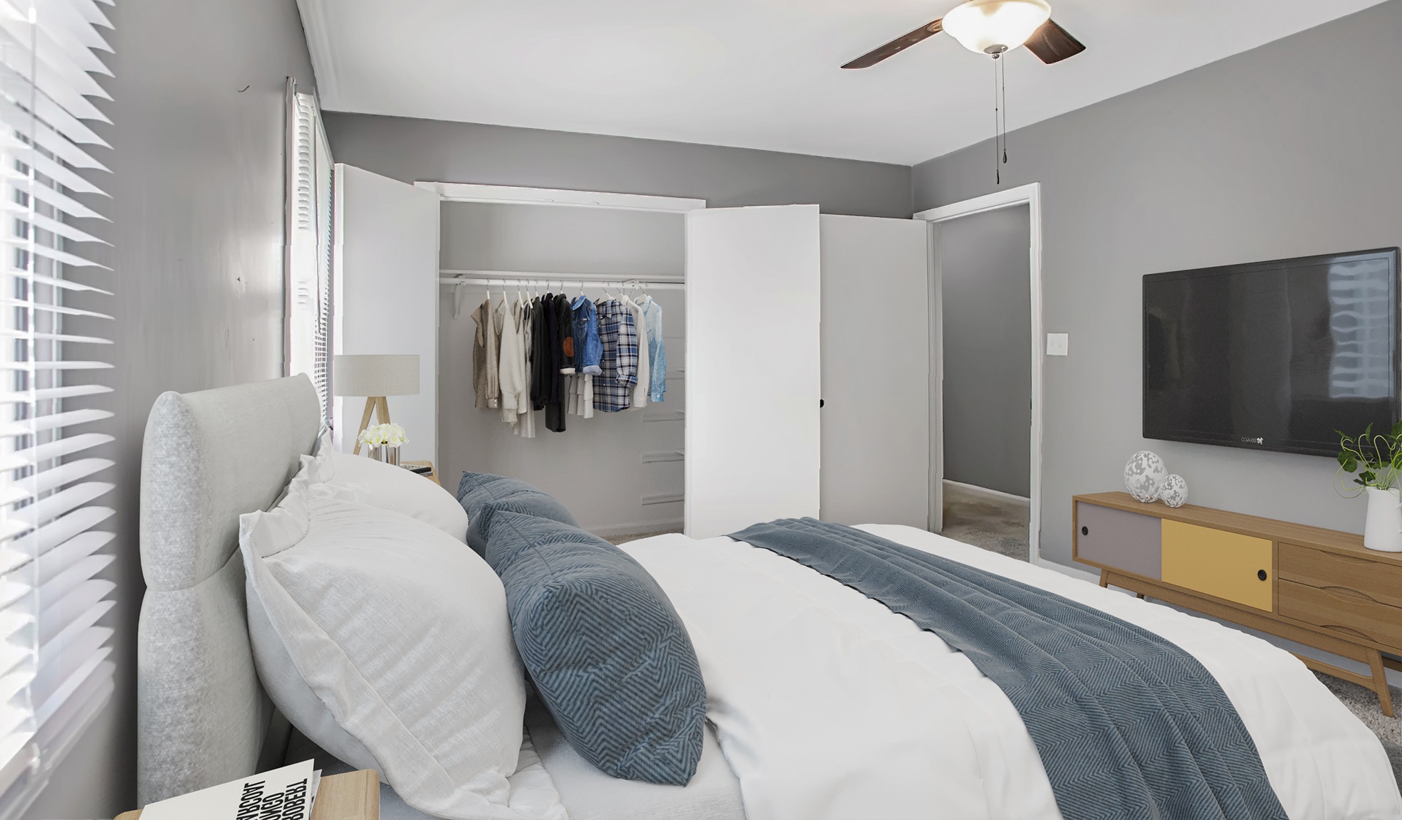 A bedroom with a bed, pillows, a ceiling fan, a mirror, a TV, and a window with blinds.